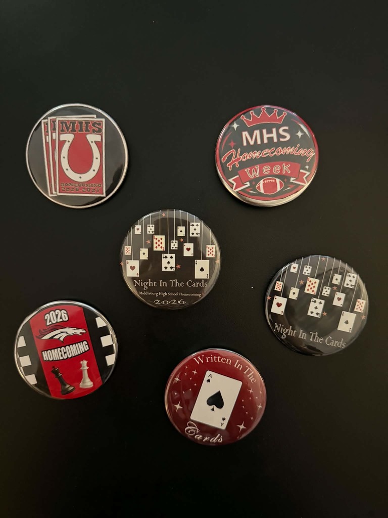 Six pins displayed on a black surface. Designs include a horseshoe, a dice set, a crown, and text like "MHS Homecoming Week".