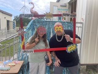 Two adults wearing superhero masks, a woman in white and a man in black, pose behind a photo frame.
