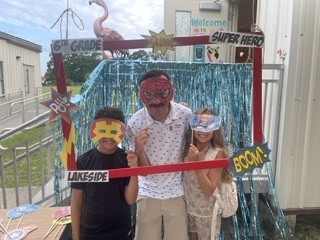 Three people stand in front of a photo frame with a blue background. One has a mask and a red cape.
