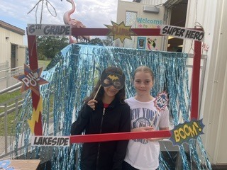 Two young girls standing in front of a photo booth with decorations reading "Lakeside" and "Super Hero."