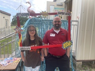 Two people stand in front of a decorated archway with "Super Hero" and "Boom!" signs. A building is in the background.