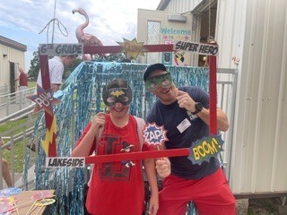 Two people pose in front of a photo booth with a superhero theme, red frame, and colorful decorations.