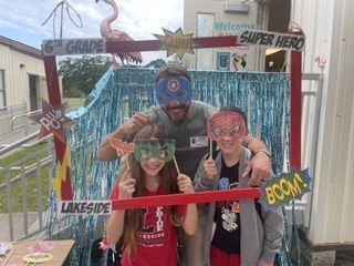 Three people pose behind a photo frame with a pink flamingo and "6th Grade Super Hero" text.