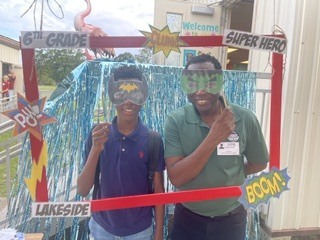Two people wearing masks pose for a photo under a sign reading "Welcome Super Hero" and "Boom!"