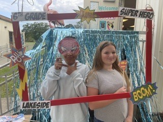 Two individuals stand in front of a backdrop with a sign reading "6th Grade Super Hero" and a pink flamingo.