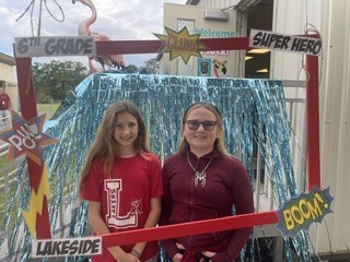 Two young girls stand behind a sign that reads "6th Grade Super Hero". They pose next to a decorative backdrop.