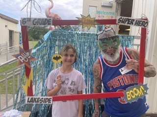 Two people stand in front of a colorful photo booth with a backdrop and sign that reads "6th Grade".