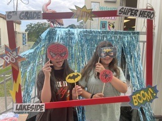 Two children in masks and capes stand in front of a decorated photo booth. One holds a stick with a Batman symbol.