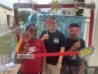 Three students stand in front of a photo booth. They wear superhero masks and hold signs. A sign reads "Welcome, 6th Grade Superhero."