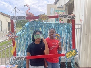 Two children, one with goggles, pose in front of a booth with a banner reading "6th Grade" and "Lakeside".