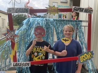 Two kids stand behind a decorated photo booth with signs reading "6th Grade" and "Super Hero".