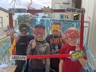 Four children wear masks with superhero labels. They stand in front of a frame with a pink flamingo and blue streamers.
