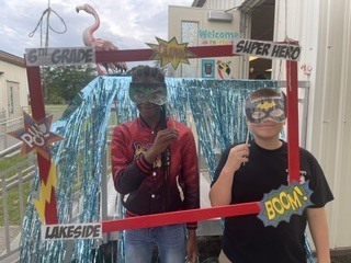 Two students in masks pose for a photo in front of a decorated frame outside a building.