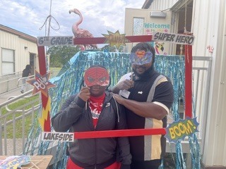 Two people stand in front of a photo booth with a red frame, wearing masks and costumes.
