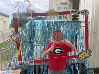 A boy in a red T-shirt and mask stands in a photo booth with blue curtains and a sign reading "Welcome Super Hero."