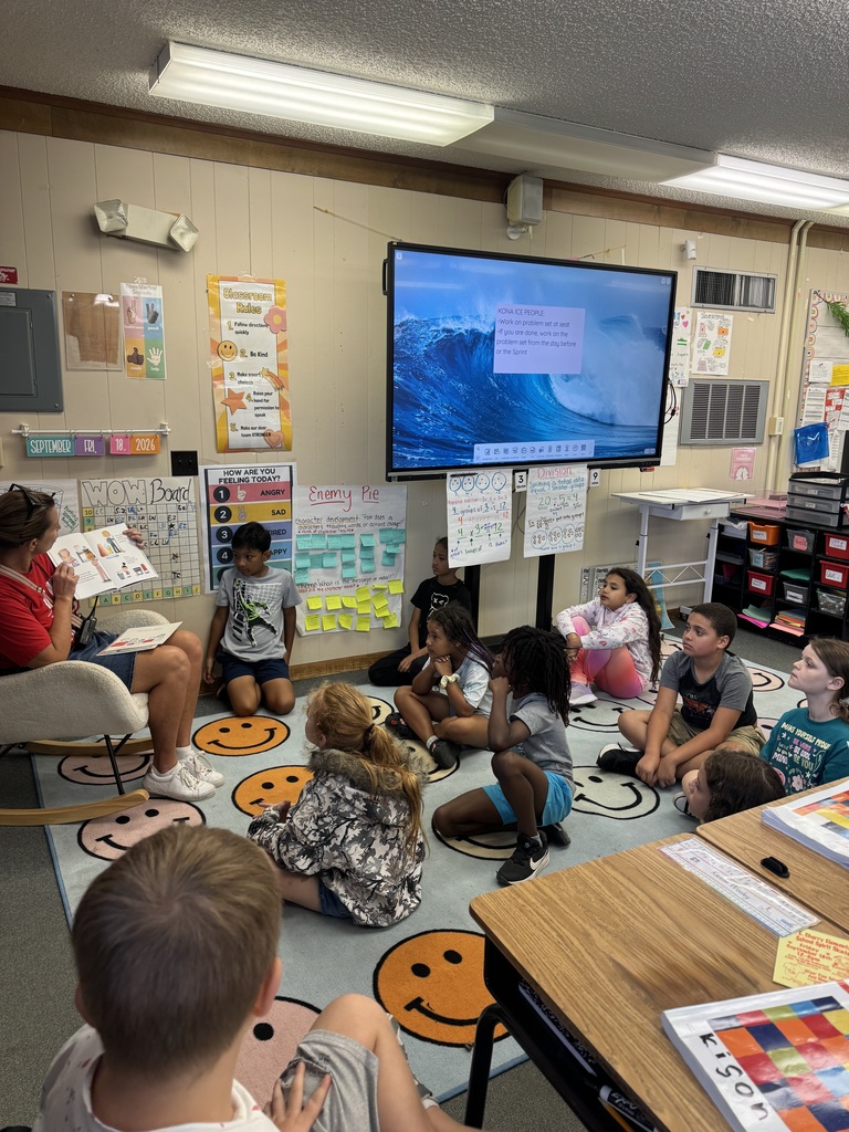 Children sit on the floor in front of a teacher. A TV screen displays a wave image.