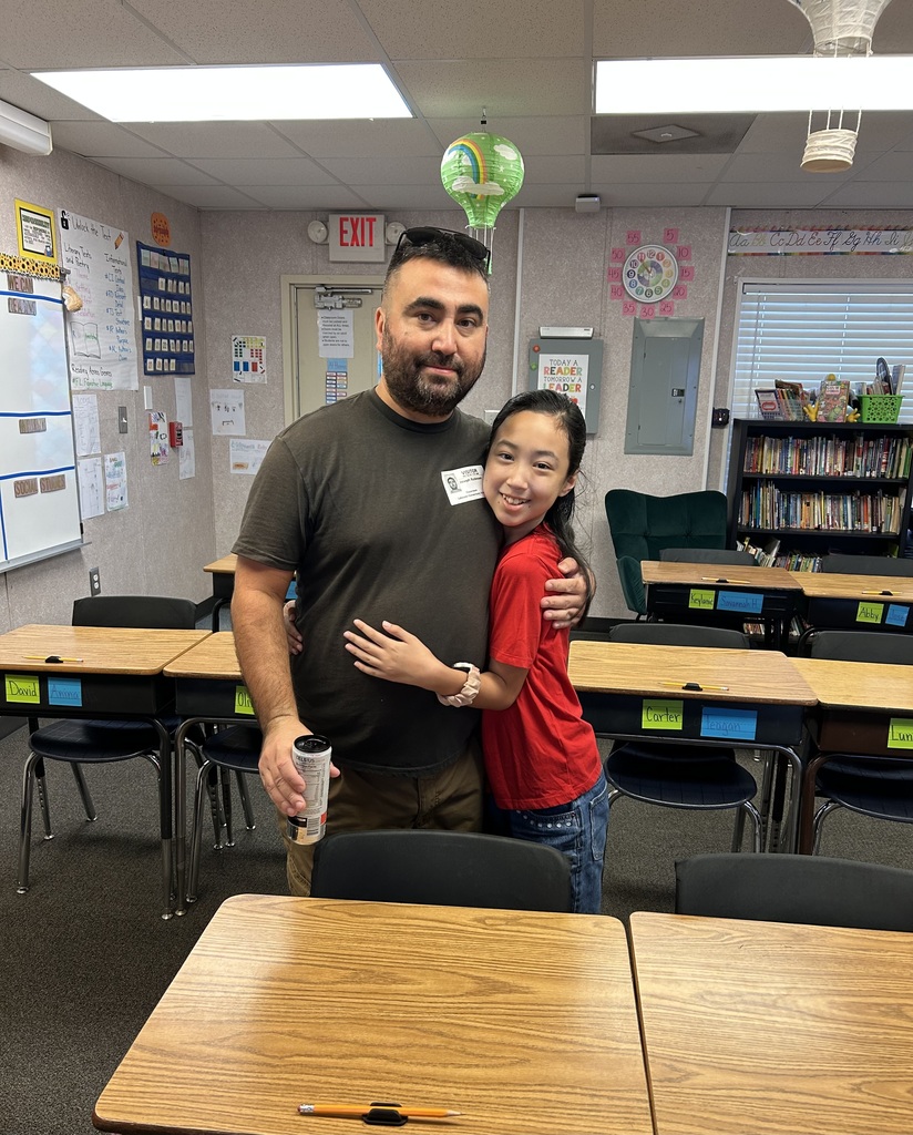 A man in a brown shirt and a young girl in a red shirt stand close, smiling. Behind them, desks and chairs are arranged in a classroom.