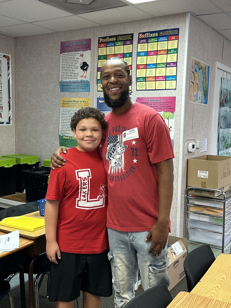 A man and a boy stand in a classroom. The boy wears a red shirt with white letters.