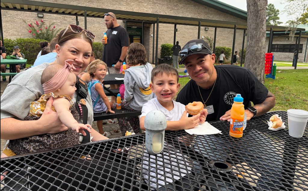 Family at a park with a baby, a boy, and a man with a donut and drinks on a table.