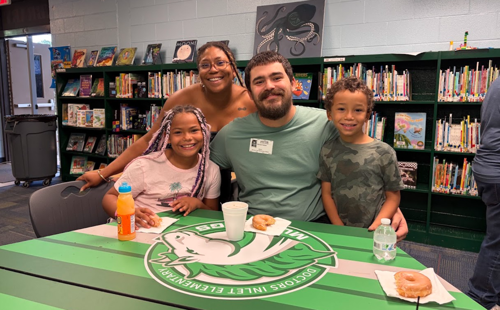 Family with two children, sitting at a table with food in a library setting with shelves in the background.