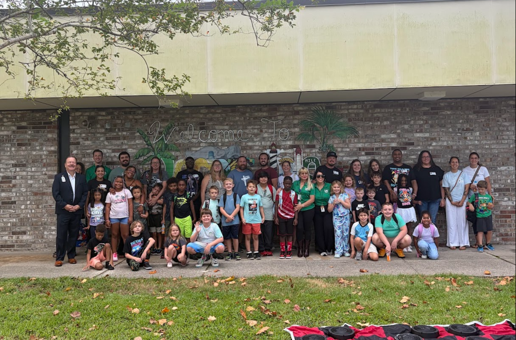 Large group of people, adults and children, standing in front of a brick wall with a mural.