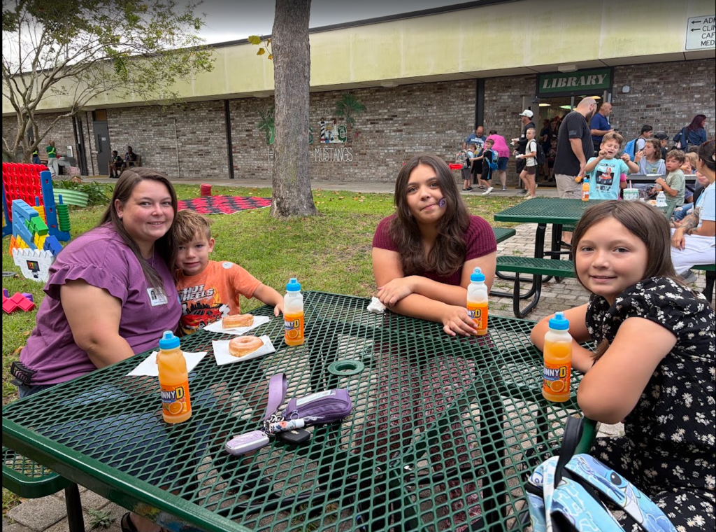 Four individuals at a park table, with food, drinks, and bags. A playground and building in the background.