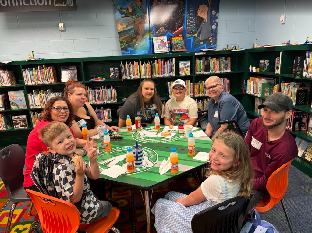 Group in a library setting, seated around a green table with drinks and snacks. Bookshelves filled with books in the background.