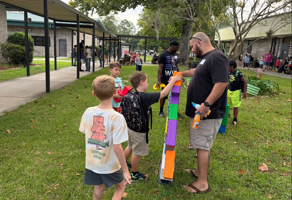 Adults and children in shorts and t-shirts stand outside. A man holds a long multicolored object.