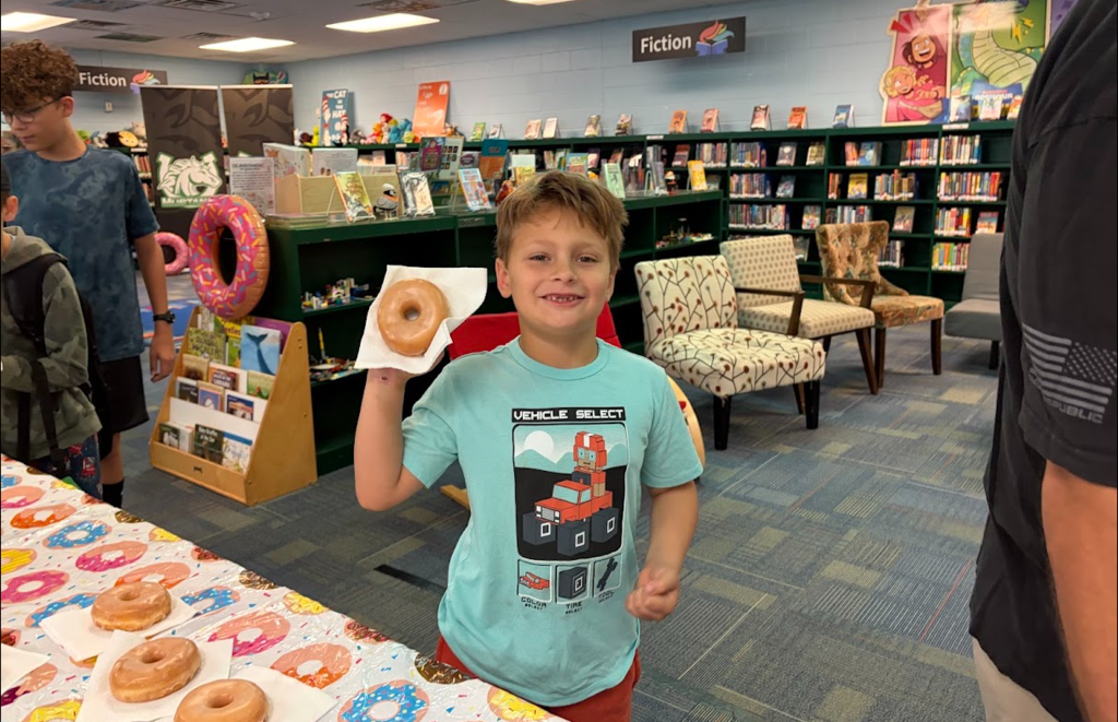 A boy in a blue shirt holds a doughnut, standing at a table with more doughnuts. Shelves with books surround him.