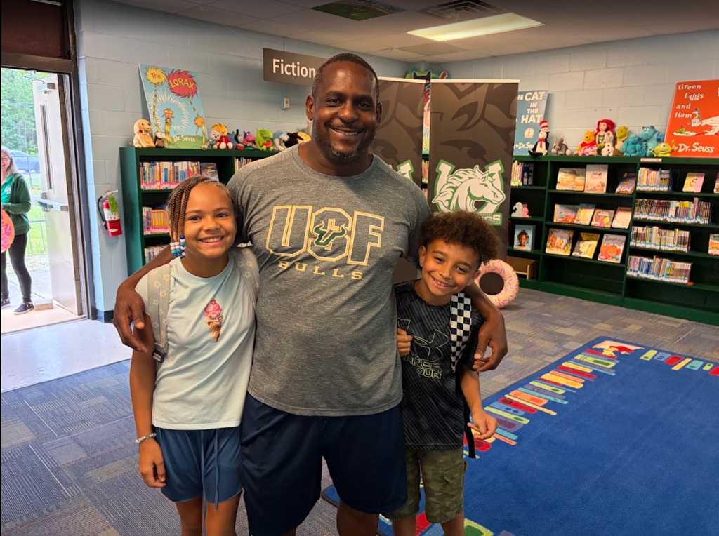 Adult man in gray T-shirt with two kids posing for a photo in a room with bookshelves and carpet.