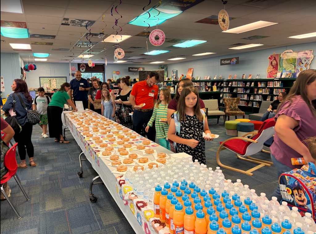 Indoor room with tables of donuts and drinks. Many children and adults stand in line.