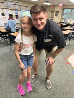 Man and young girl standing in a classroom with desks and chairs in the background.