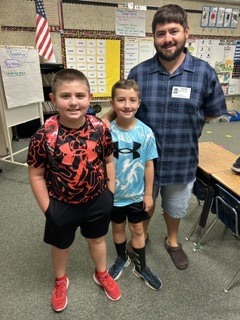 Three children in casual clothes pose in a classroom. A flag hangs in the background.