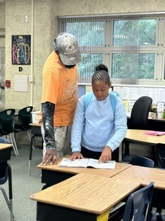 Adult and child in a classroom, standing at a desk, looking at a book. Behind them are chairs and windows.