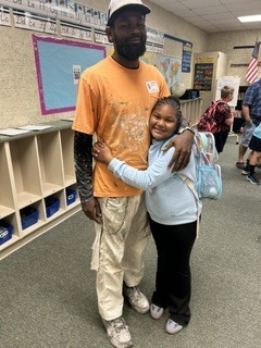 A man and a girl stand close, smiling. The man wears an orange shirt and pants. The girl hugs him with a backpack. Behind them, shelves, a bulletin board, and people are present.