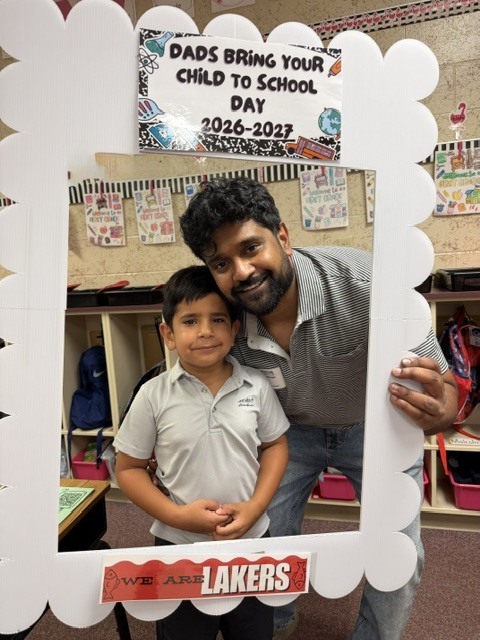 Man and young boy in a white frame that says, "Dads Bring Your Child to School Day 2026-2027."