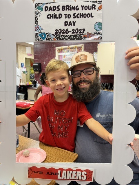 A father and son hold a photo frame reading "Dads Bring Your Child to School Day 2026-2027" with a Lakers logo in a classroom.
