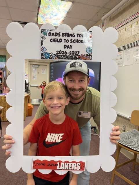 A man and a boy in front of a frame. The boy has a Nike shirt. Behind them is a white wall with a monitor.