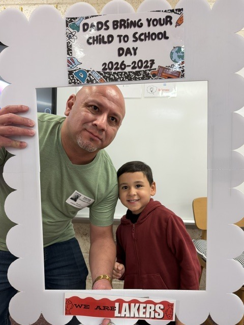 A man holds a photo frame with a boy in front of a sign that reads "Dads Bring Your Child to School Day 2026-2027".