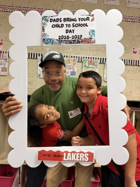 A man wearing a green shirt and cap stands with two children. They pose in a white frame that says "Dads Bring Your Child to School Day".