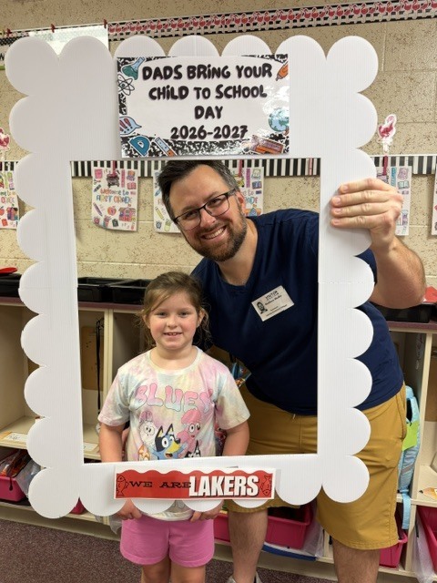 A man and a young girl stand before a frame with "Dads Bring Your Child to School Day" printed on it.