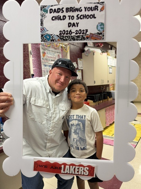 A man and a boy stand in a white frame with a message reading "Dads Bring Your Child to School Day 2026-2027."