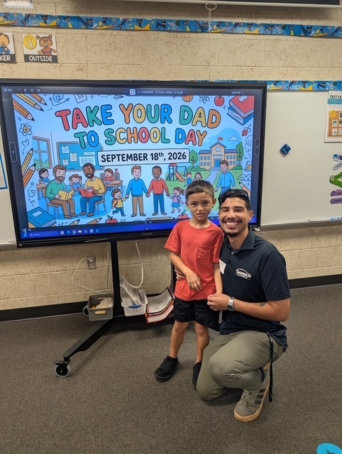 A father and son pose in a classroom. Behind them is a monitor with a Take Your Dad to School Day graphic.