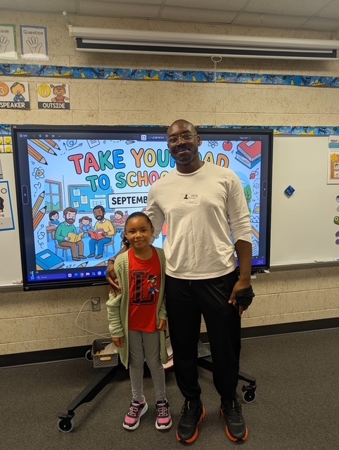 A man and a child standing next to each other in a classroom, in front of a monitor.