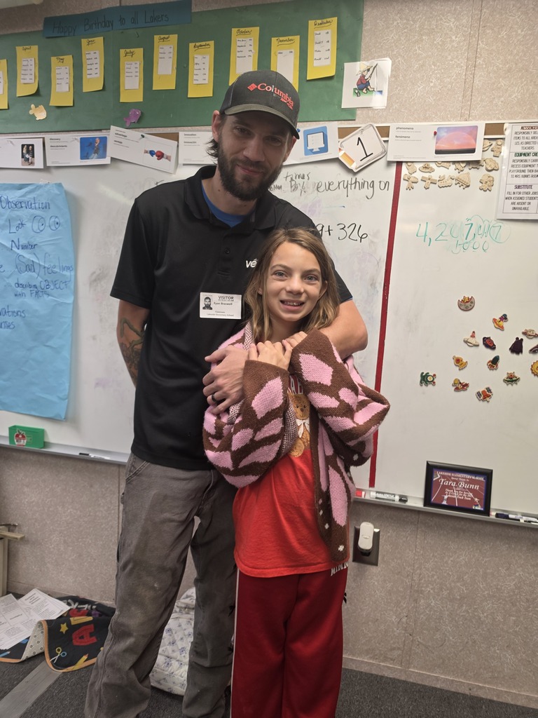 A man in a black shirt and cap stands with a young girl wearing a pink shirt and red pants. They are in front of a whiteboard with various decorations.