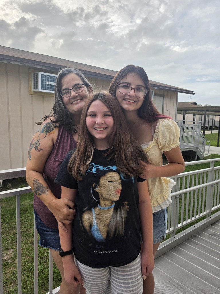 Three women stand on a porch. The girl in the middle wears a black t-shirt. Behind them, a house is visible with a metal railing.