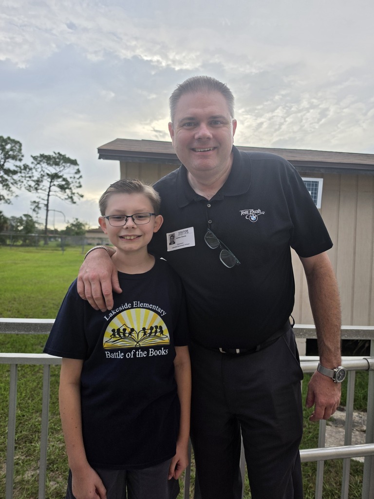 A man and a boy stand together in front of a fence, smiling. The man wears a black shirt, and the boy wears glasses and a yellow t-shirt.