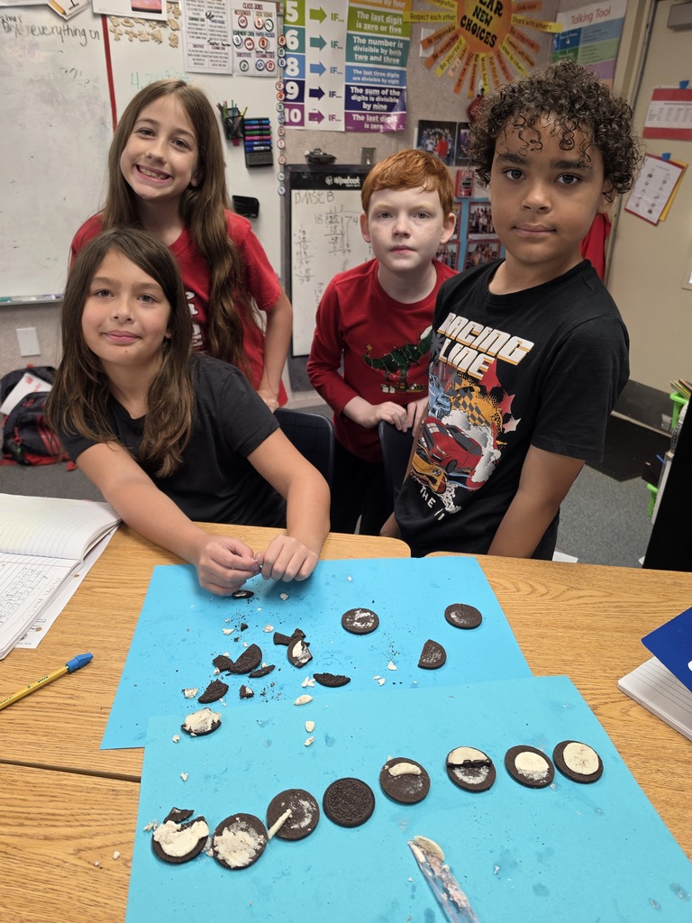 Four children in a classroom with a table covered in blue paper, displaying cookies and cookie pieces.