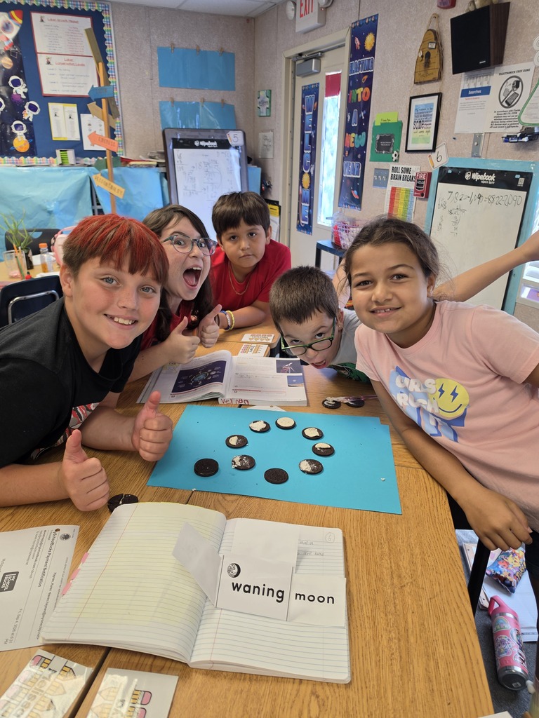 Five kids at a table with a blue board and game pieces, open book, and classroom walls in the background.