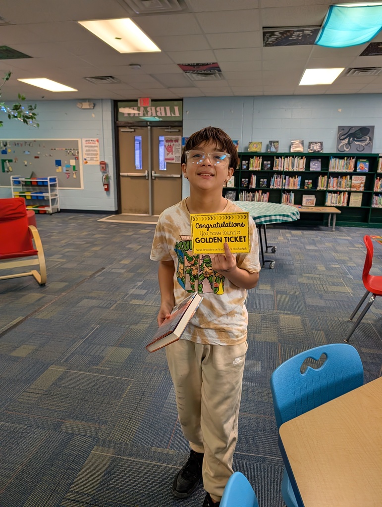 A boy stands in a library holding a book and a yellow card. He wears glasses, sneakers, and a t-shirt. Behind him, there are chairs, tables, a fire extinguisher, and a bulletin board.